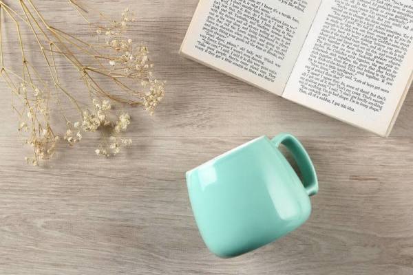 Açık ahşap bir yüzey üzerinde yatık duran mavi bir fincan, yanında kurutulmuş çiçekler ve açık bir kitap.