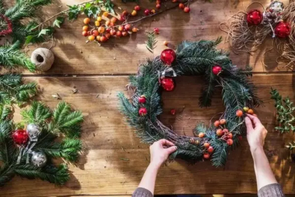 Bir kişi, çam dalları ve süslemelerle dolu bir masada Noel çelengi yaparken.