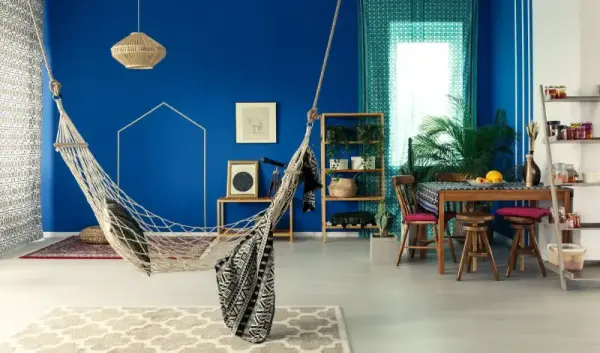 Mavi duvarlı, modern bir oda içinde hamak, yemek masası ve süs eşyaları.