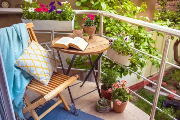 Balkonda ahşap sandalye, yastık, mavi battaniye, kitap ve çeşitli saksı bitkileri.