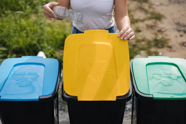 Bir kişi, üç farklı renkte kapaklı geri dönüşüm kutularının önünde plastik bir şişe tutuyor.