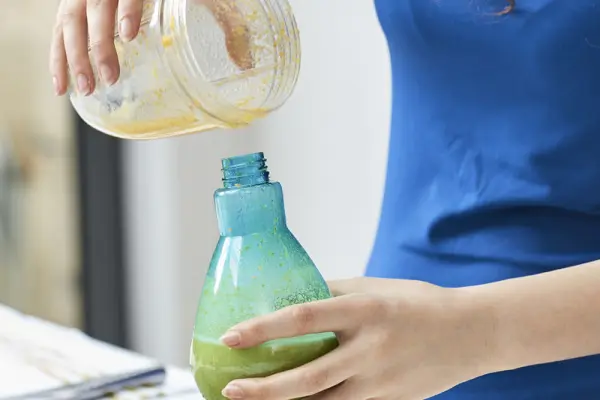 Mavi önlüklü bir kişi, kalan deterjanı eski bir plastik şişeden yeni bir şişeye aktarıyor.