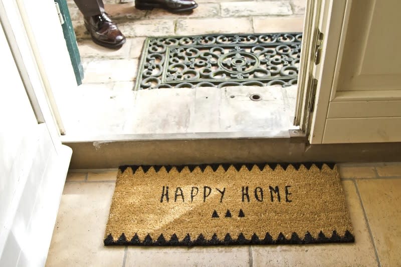 Eve girişte "Happy Home" yazılı paspas, kapı açık, içeride insan ayakları görülüyor.