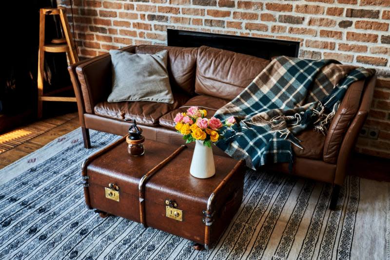 Kahverengi deri kanepe, tuğla duvar, vintage bavul sehpa üzerinde çiçekler ve kilim.