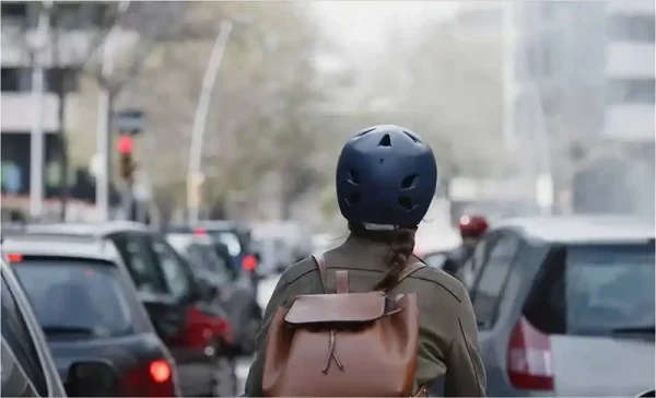 Bisiklet kaskı takmış ve sırt çantası bulunan bir kişi, trafikteki araçlara arkadan bakıyor.