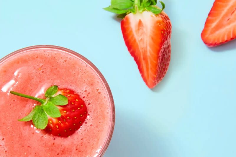 Bir bardak çilek smoothie'si ve iki yarım çilek, açık mavi arka planda.