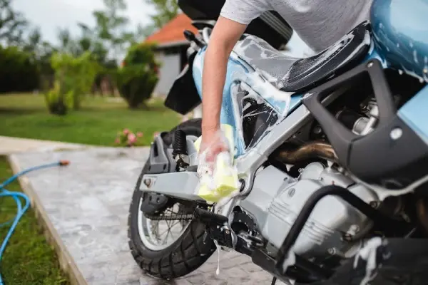 Bir kişi, bir bahçede park halindeki mavi bir motosikleti sünger ile yıkıyor.