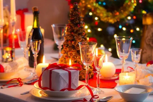 Yemek masası üzerinde mumlar, hediyeler ve arkada renkli ışıklarla süslenmiş bir Noel ağacı.