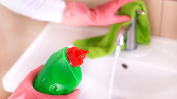 Temizlik yapmak için eldiven takmış bir kişi, yeşil bir bez ile lavabo temizliyor ve elinde yeşil bir deterjan şişesi tutuyor.